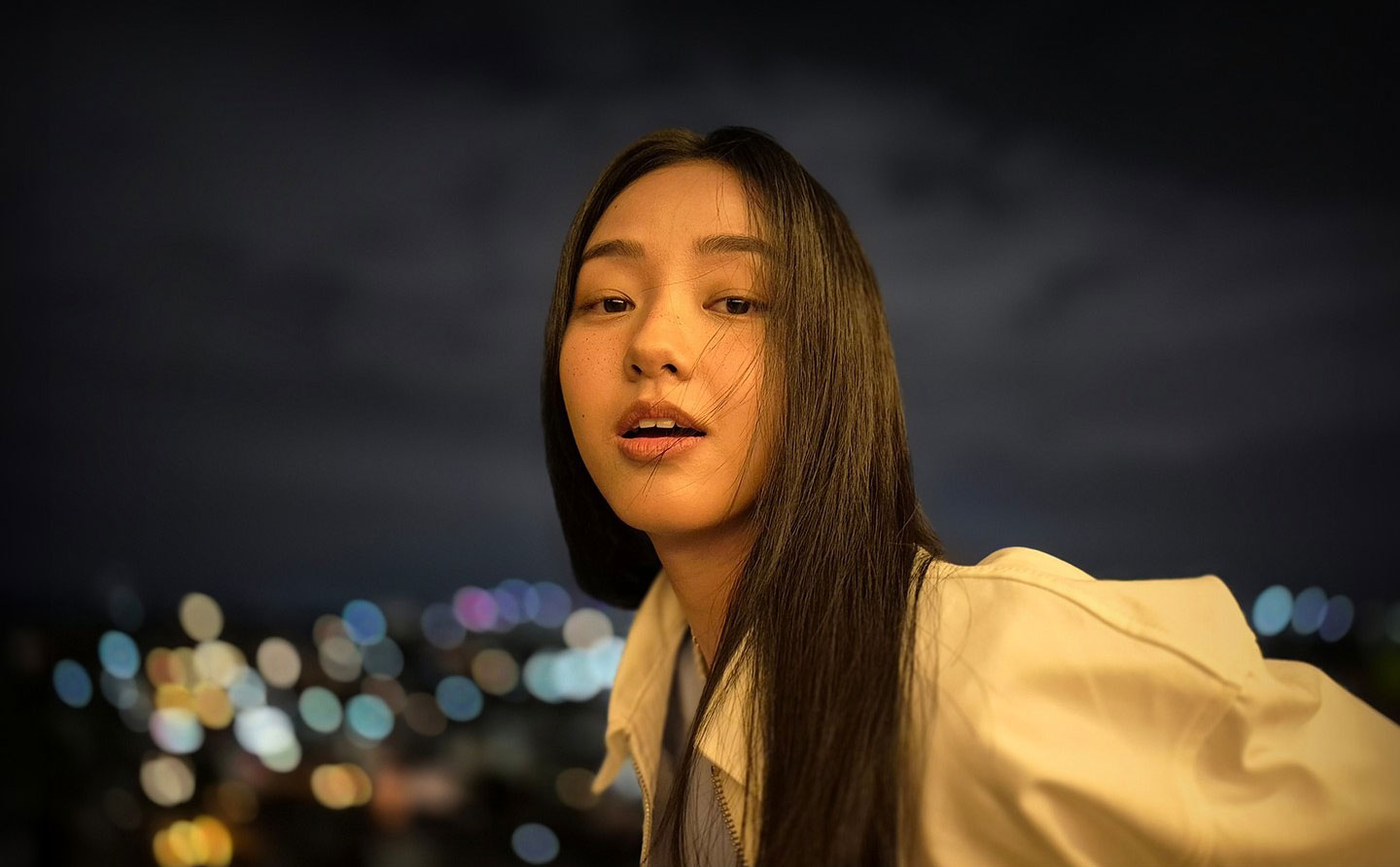 A photo of a woman outdoors at night with a lit face and blurred city lights in the background. Text reads Captured by Galaxy S24 FE #withGalaxy.